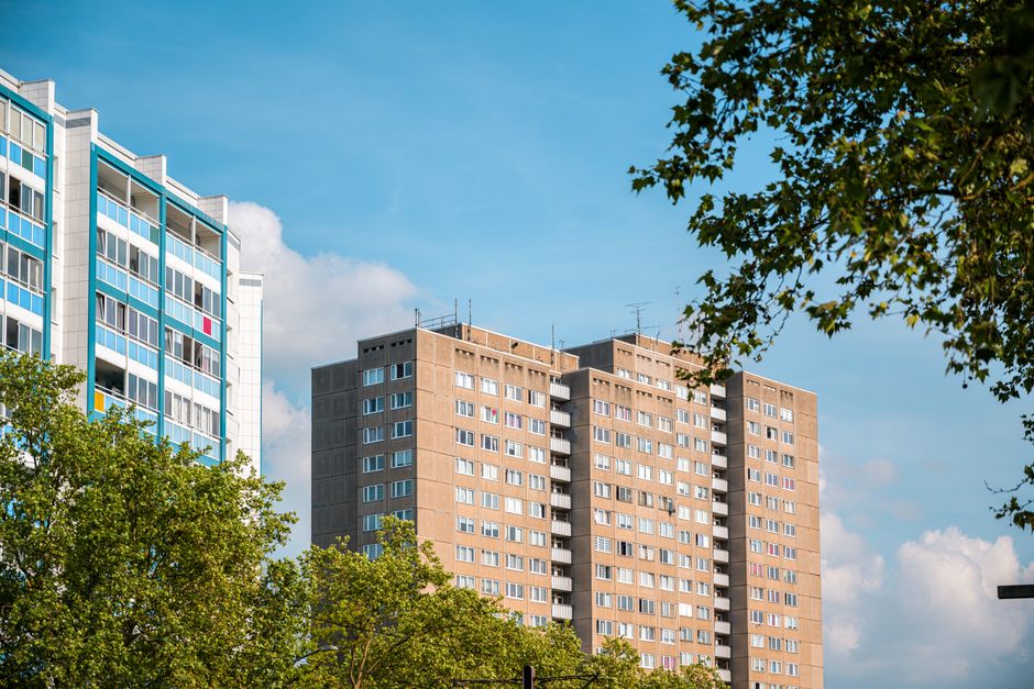 A low-angle view of two tall residential apartment buildings with numerous windows, some of which have opened or closed blinds. The building on the left features prominent yellow horizontal panels acr